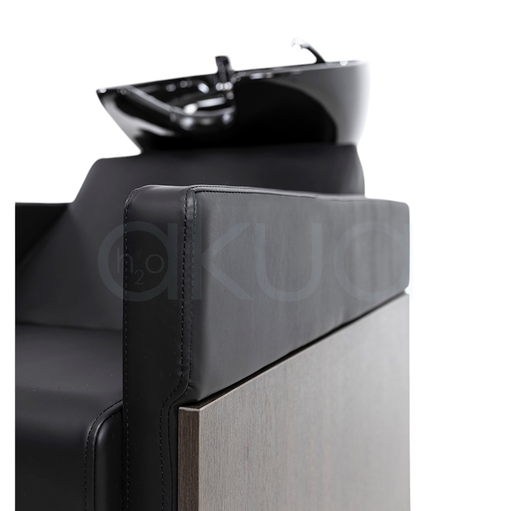 Lavacabezas Rustik negro H2oAkua. La imagen muestra un lavacabezas de peluquería moderno, compuesto por un sillón tapizado en color negro con apoyabrazos anchos y estructura lateral de aspecto madera. En la parte superior, integrado al respaldo, se encuentra una cubeta o pila de lavado de cerámica negra diseñada para lavar el cabello de los clientes.