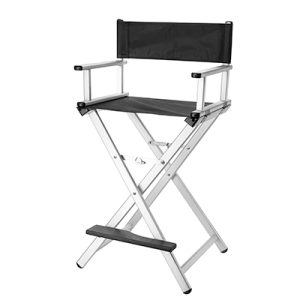 Silla maquillador plata 06156/54. La imagen muestra una silla plegable de director plata tipo taburete alto profesional, vista de frente y ligeramente de lado sobre fondo blanco. Tiene estructura tubular de aluminio negro mate con diseño en tijera para plegado compacto, asiento y respaldo rectangulares tapizados en tela negra transpirable resistente, reposapiés metálico transversal y altura ajustable para uso en salones de peluquería, maquillaje o barberías. H2o Akua