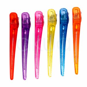 Pinza toga grande paquete 6 unid plastico metal Eurostil. La imagen muestra un conjunto de 6 pinzas de peluquería tipo clip, alineadas sobre un fondo blanco. Están fabricadas en plástico translúcido de colores vivos: rojo, morado, rosa, amarillo, azul y naranja. Cada pinza tiene un diseño alargado y estrecho, con forma ligeramente puntiaguda en la parte inferior. En la zona superior se aprecia el mecanismo metálico con muelle, que permite abrir y cerrar la pinza. El cuerpo presenta una textura o relieve antideslizante, pensado para mejorar el agarre del cabello. H2o Akua