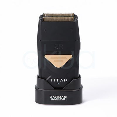 Afeitadora Titan Ragnar. Afeitadora profesional de acabado tipo foil en color negro mate con detalles metálicos dorados. En la parte superior incorpora un doble cabezal de láminas metálicas diseñado para proporcionar un afeitado preciso y apurado. El cuerpo presenta un diseño compacto y ergonómico con un panel triangular dorado en la zona frontal y la inscripción "TITAN" en letras blancas. La afeitadora está colocada sobre una base de carga negra con el logotipo "Ragnar Barber Line" visible en la parte frontal. La imagen se presenta sobre un fondo claro, resaltando el aspecto moderno, elegante y profesional de la herramienta, especialmente orientada a barberías y acabados de precisión. H2o Akua