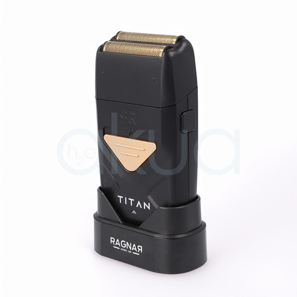 Afeitadora Titan Ragnar. Afeitadora profesional de acabado tipo foil en color negro mate con detalles metálicos dorados. En la parte superior incorpora un doble cabezal de láminas metálicas diseñado para proporcionar un afeitado preciso y apurado. El cuerpo presenta un diseño compacto y ergonómico con un panel triangular dorado en la zona frontal y la inscripción "TITAN" en letras blancas. La afeitadora está colocada sobre una base de carga negra con el logotipo "Ragnar Barber Line" visible en la parte frontal. La imagen se presenta sobre un fondo claro, resaltando el aspecto moderno, elegante y profesional de la herramienta, especialmente orientada a barberías y acabados de precisión. H2o Akua